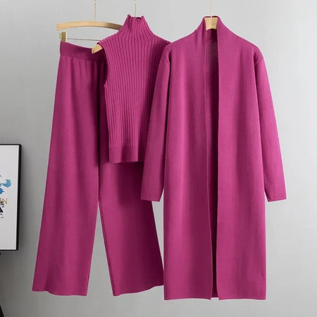 A pink three-piece women's coordinated outfit consisting of a long cape, a pair of wide-leg pants, and a high-neck top, displayed on hangers against a grey background.