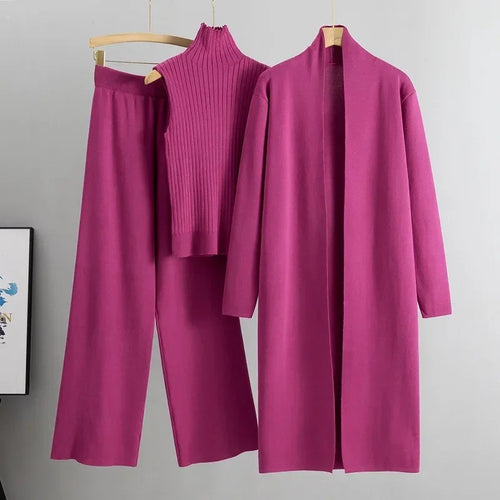 A pink three-piece women's coordinated outfit consisting of a long cape, a pair of wide-leg pants, and a high-neck top, displayed on hangers against a grey background.